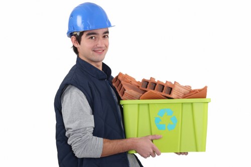 A young man wearing a blue safety helmet, gray long-sleeved shirt, and navy vest is smiling while holding a green recycling bin with a blue recycling symbol on the side. The bin contains a stack of red bricks, indicating construction or renovation debris. The background is plain white, emphasizing the individual and the waste container, which is likely part of a rubbish removal service for domestic or commercial property clearances around Wimbledon. The image highlights professional waste management practices, with the individual preparing to transport the debris for proper disposal or recycling, fitting within the context of rubbish removal services offered by Office Clearance Wimbledon in SW19 postcode area. The containers and bricks suggest the handling of building waste commonly associated with office or property clearance in the local area, reflecting the company's focus on responsible waste management and environmental consciousness.