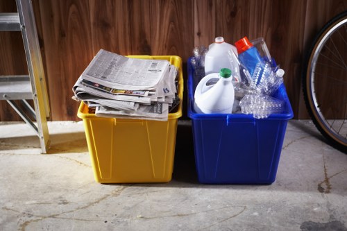 Two large plastic recycling bins placed on a concrete floor in an indoor or garage area with wooden paneling in the background. The yellow bin on the left is filled with a stack of newspapers and magazines, while the blue bin on the right contains clear plastic bottles, a white plastic jug, and some empty plastic containers, suggesting they are used for recycling waste materials. To the right of the bins, part of a bicycle wheel is visible, indicating the location might be a residential garage or storage space. The lighting is natural or from an overhead source, casting soft shadows and highlighting the textures of the plastic and paper. This setup reflects usual rubbish and recycling collection practices, potentially related to local waste removal services in Wimbledon or nearby areas, as part of general rubbish disposal efforts in the region.
