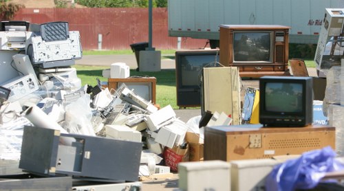 A large collection of discarded electronic waste, including old CRT televisions, computer monitors, VCR players, and various electronic components, is piled outdoors on a paved surface. The devices display various colours and screen types, with some showing reflective screens and others with wooden or plastic casings. The background includes a grassy area, a red fence, and some trees, indicating the waste is situated in an outdoor environment near a residential or urban area. The electronic items are stacked and somewhat scattered, with some screens facing different directions and components overlapping. The overall scene suggests a collection of outdated or broken electronic devices awaiting disposal or recycling, typical for rubbish removal services in Wimbledon or nearby areas. Office Clearance Wimbledon undertakes the removal and proper disposal of such electronic waste as part of their rubbish clearance services to ensure environmentally responsible recycling and clearance from the property, which might be in the Wimbeldon area postcode.