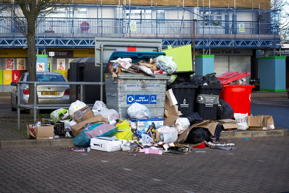 An overflowing urban waste collection point situated on a paved street, featuring a large grey mixed paper and cardboard recycling bin with a partially open lid revealing crumpled paper, cardboard box