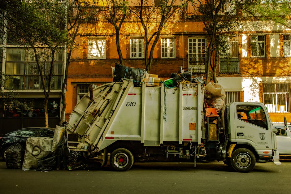 A worker wearing a high-visibility yellow vest and dark clothing stands next to a large white rubbish collection vehicle on a city street during dusk. The vehicle features red and orange reflective ch